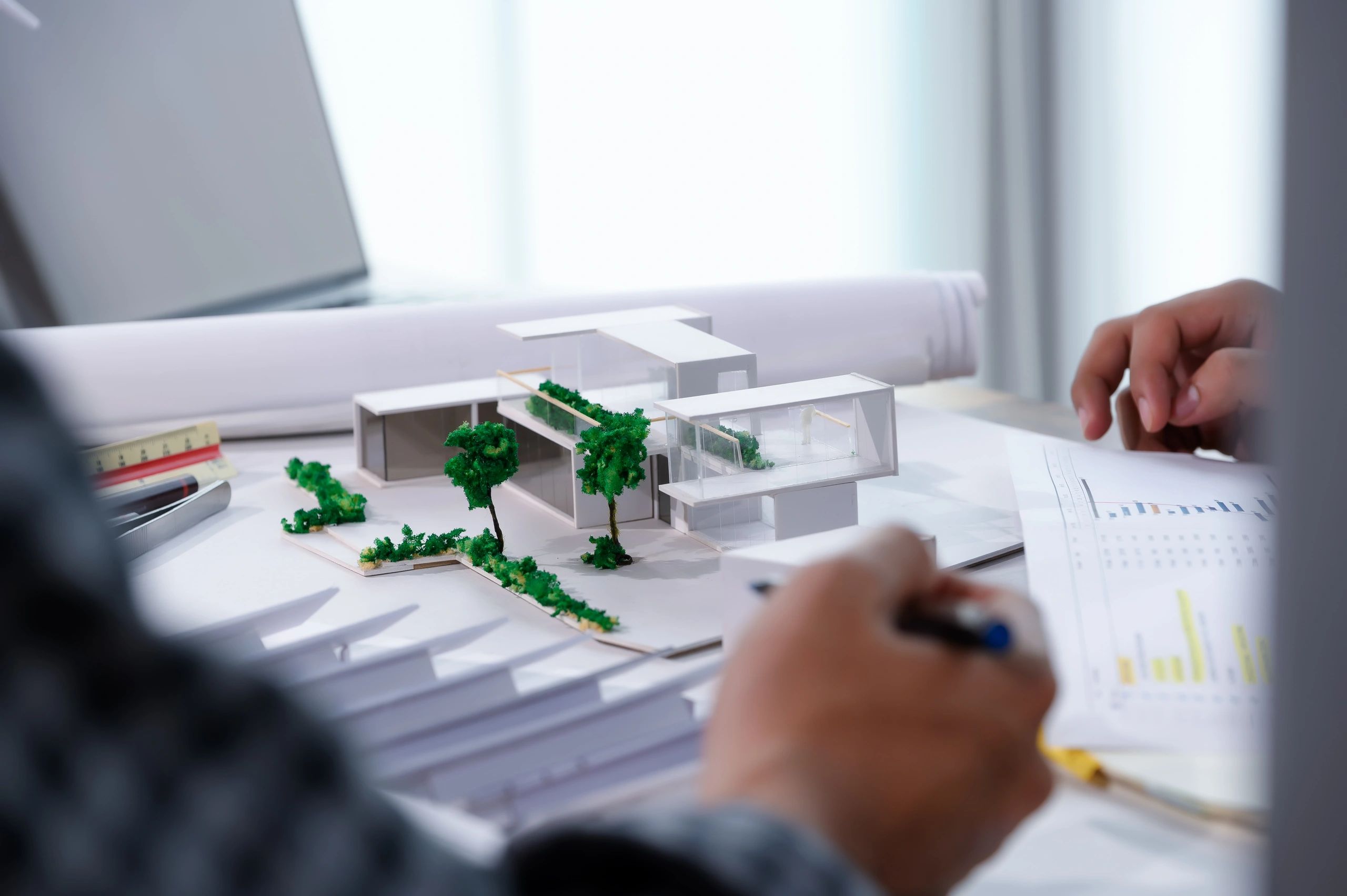 A person examines a building model with greenery on a desk, surrounded by architectural plans and documents.